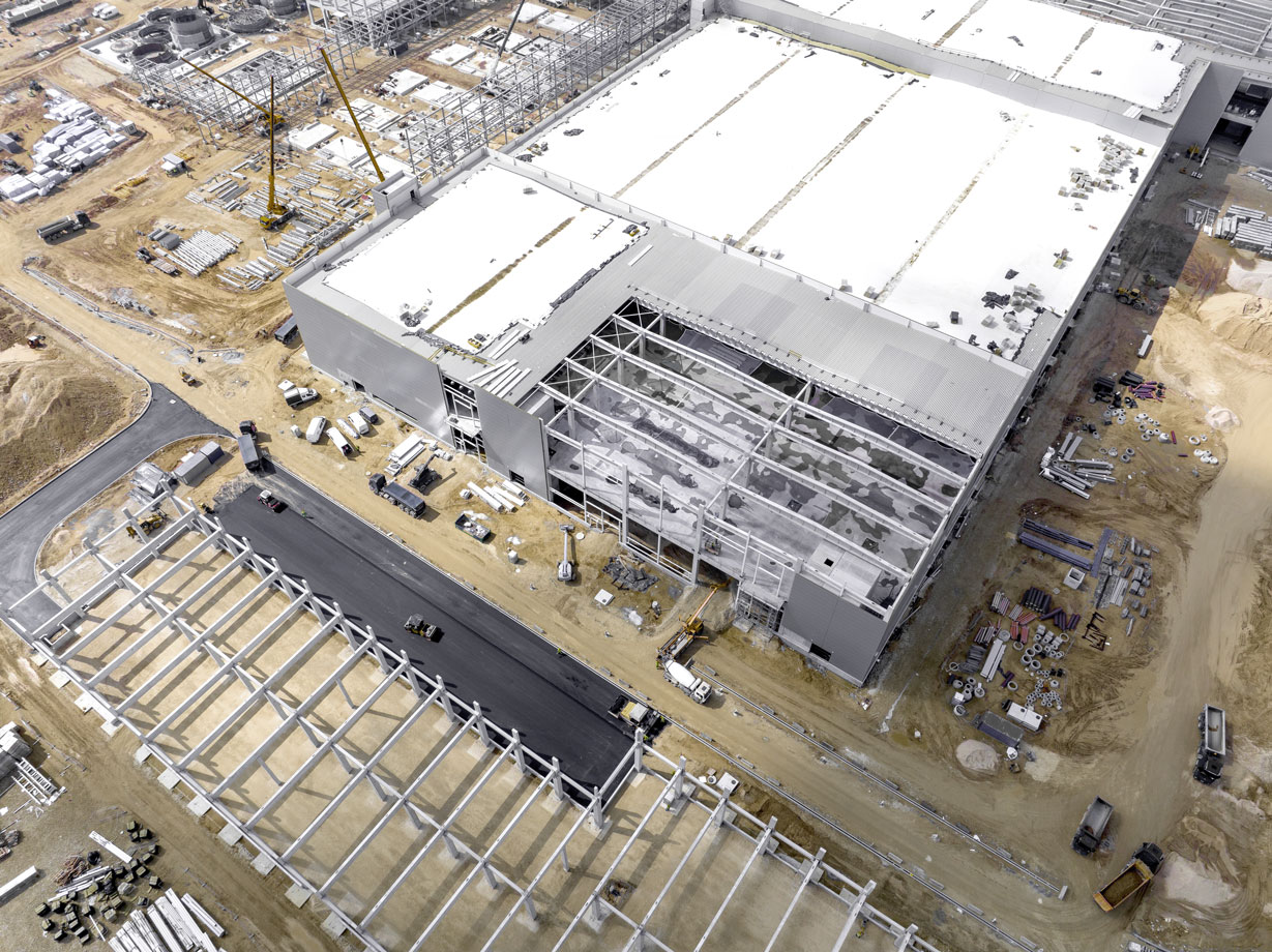 Luftaufnahme einer Baustelle mit teilweise errichtetem Industriegebäude und Stahlrahmenkonstruktion auf sandigem Gelände